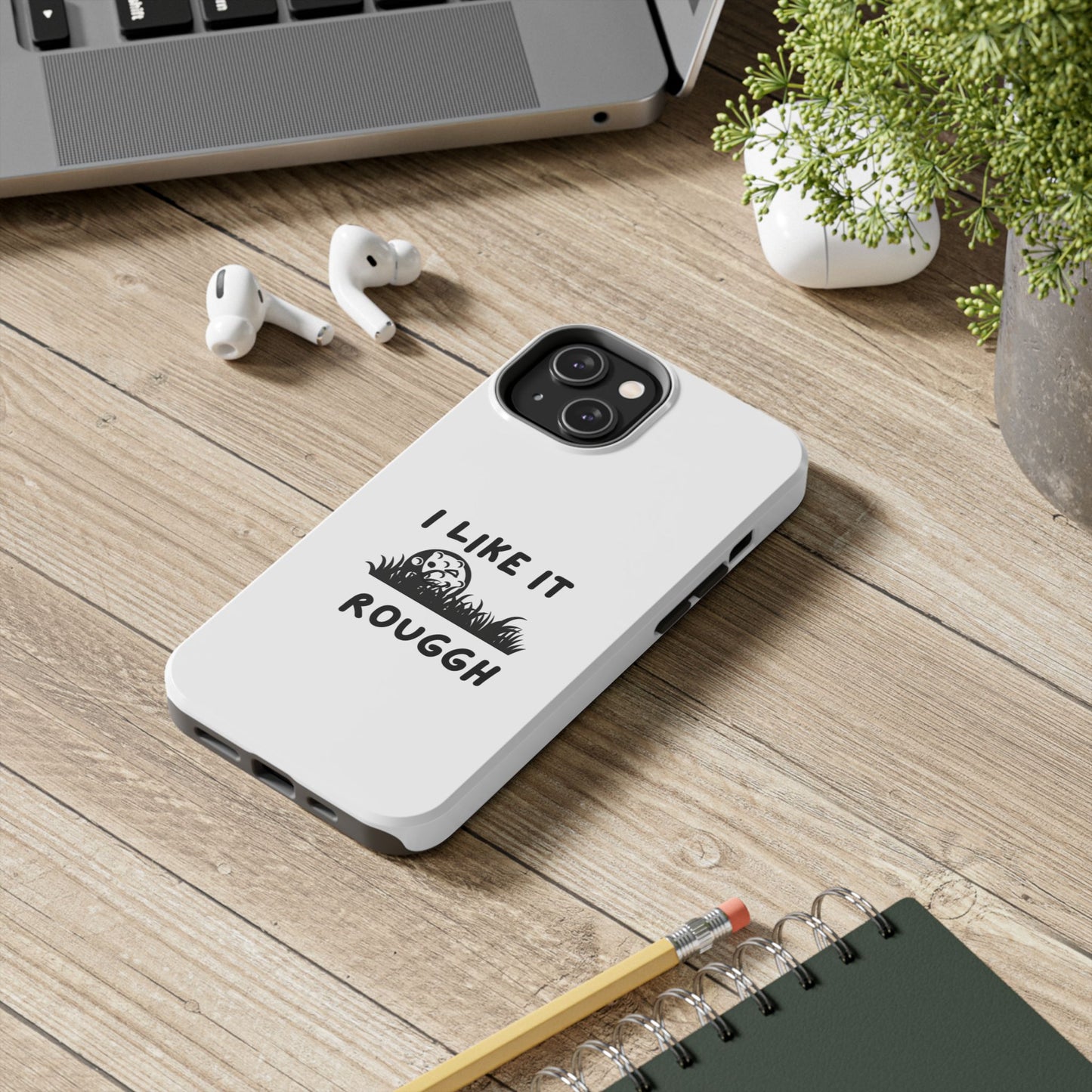 Phone case with 'I Like It Rough' text on a wooden desk with laptop, earbuds, and notebook.